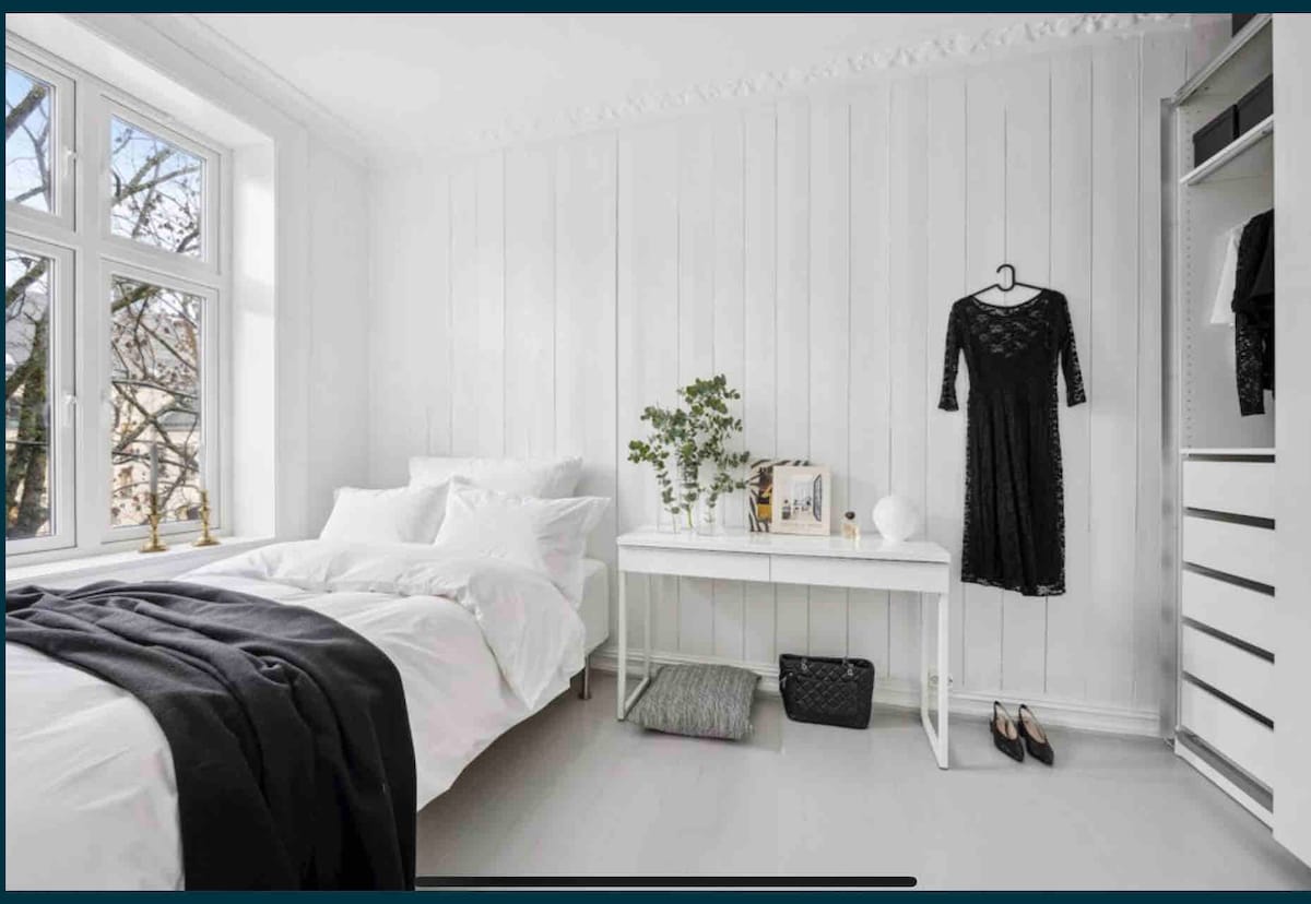 A light-filled bedroom features white paneled walls and a spacious bed dressed in soft linens. A black dress hangs on a hook beside a minimalist table holding decorative items. Natural light filters through large windows, illuminating a cozy and inviting atmosphere.