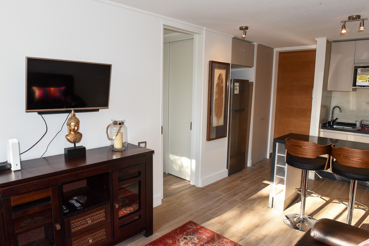 A modern living area features a flat-screen TV mounted above a wooden cabinet. A dining counter with high stools sits adjacent to an open kitchen, displaying contemporary appliances. Natural light streams through a nearby doorway, highlighting the warm wood flooring.