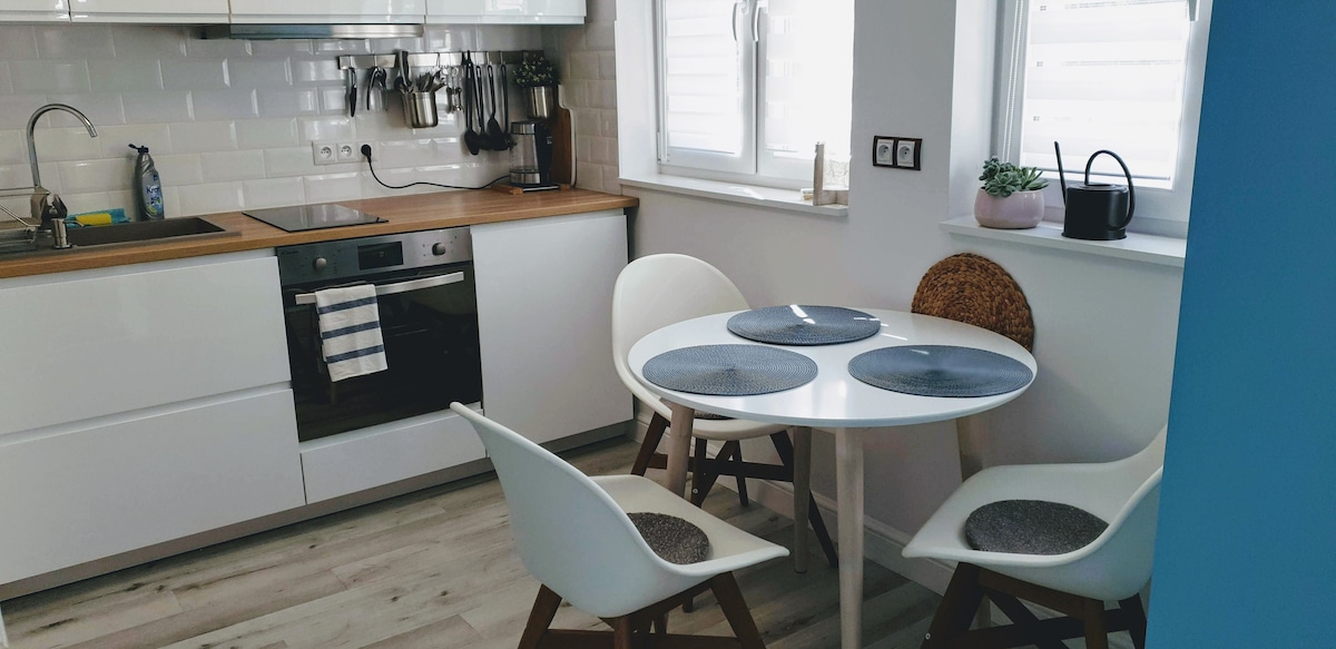 A bright kitchen area features modern white cabinetry and a wooden countertop. A small round table is surrounded by four chairs, including two with woven seats. Natural light enters through two windows, with a small plant situated on the windowsill.