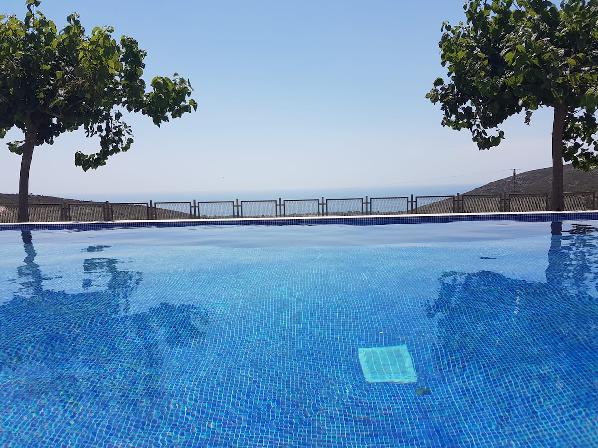 A clear swimming pool reflects the sky, surrounded by lush trees. The pool offers unobstructed views of the sea and distant hills, with a fence visible in the background outlining the property boundary.