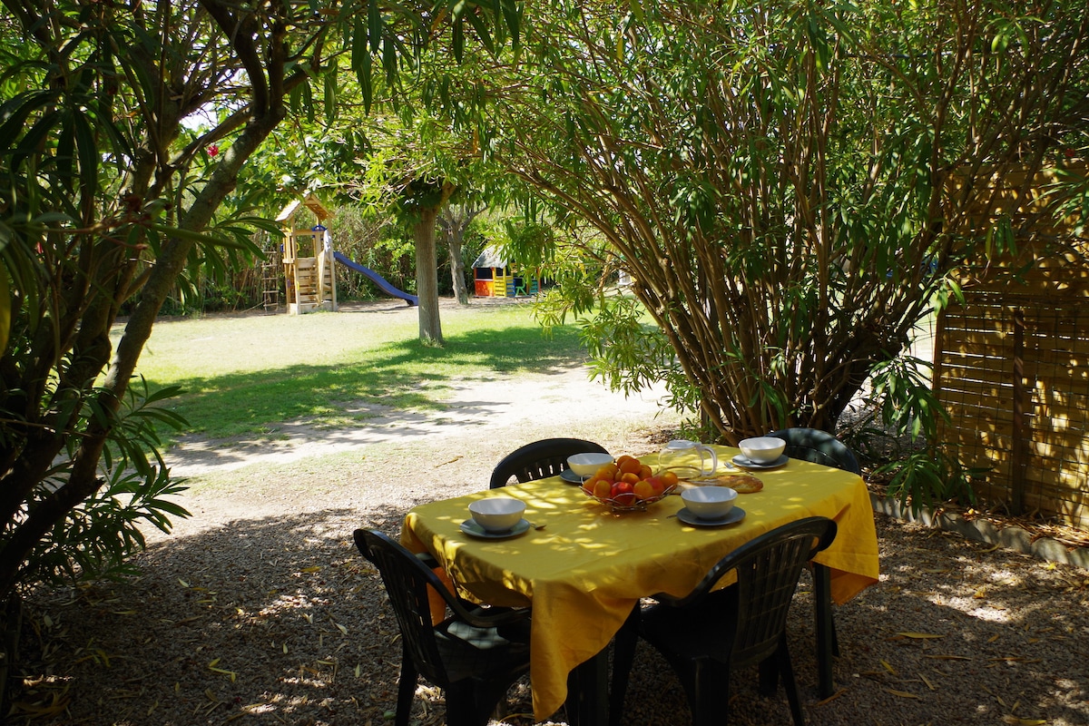 A shaded dining area features a yellow tablecloth with a set table, including bowls and a centerpiece of fruit. In the background, a playground structure is partially visible along with an expansive green lawn, framed by lush trees providing a tranquil setting.