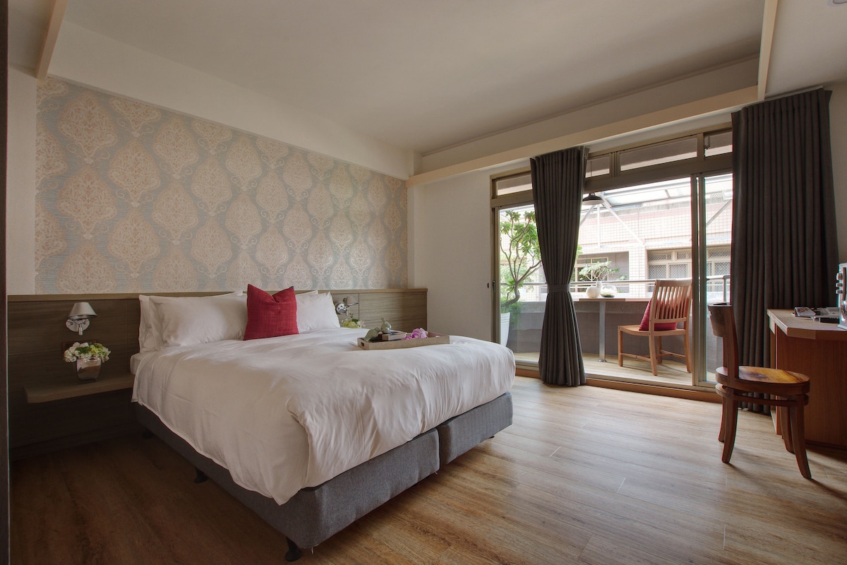 A well-lit bedroom features a spacious bed with a white comforter and decorative pillows. A large window provides natural light and a view of a balcony furnished with wooden chairs. Light wood flooring complements the soft wall decor.