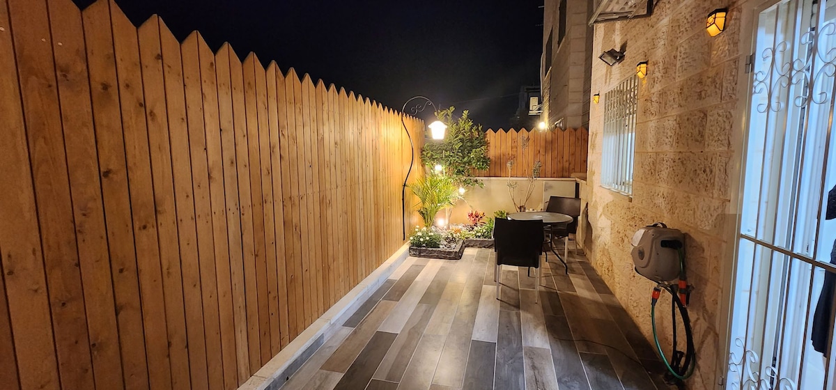 A private outdoor space features a wooden fence providing a sense of enclosure. Potted plants are seen among decorative elements, while a round table and chairs offer a cozy seating area. Soft lighting fixtures illuminate the area, creating a welcoming ambiance.