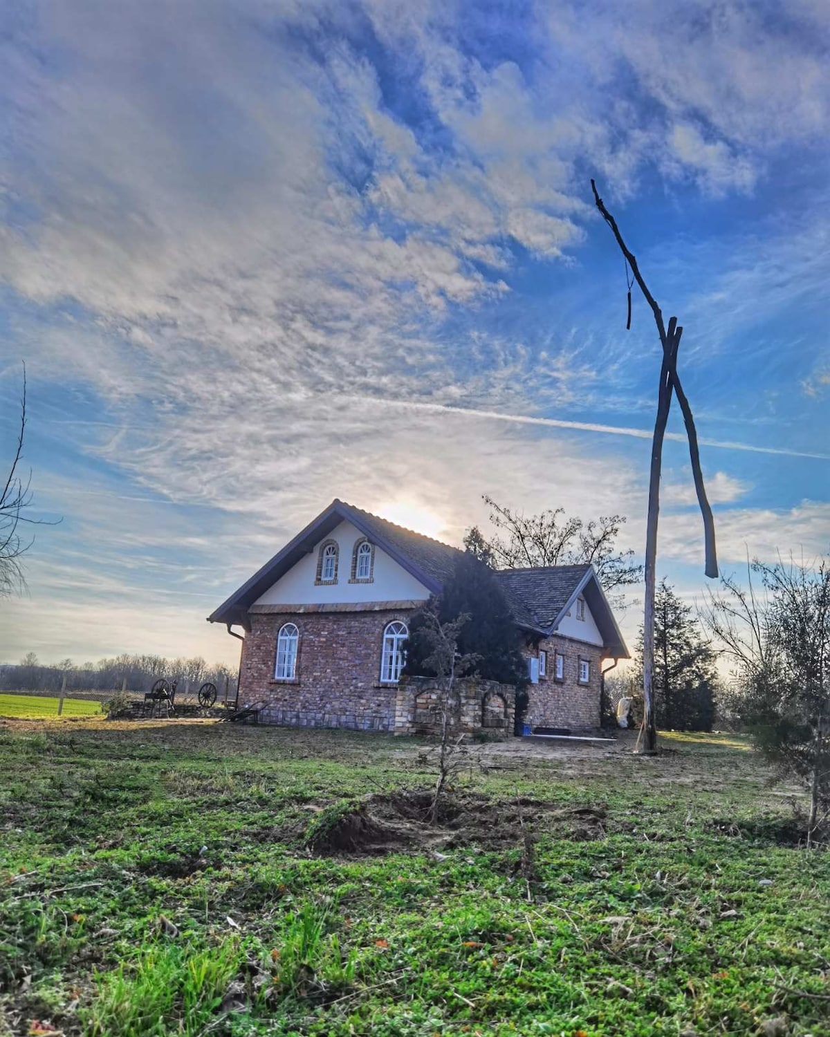 A rustic farmhouse is situated in a serene rural setting, surrounded by lush greenery. The structure features stone walls and arched windows, complemented by an expansive sky and gentle clouds. Nearby trees and open fields enhance the peaceful ambiance.