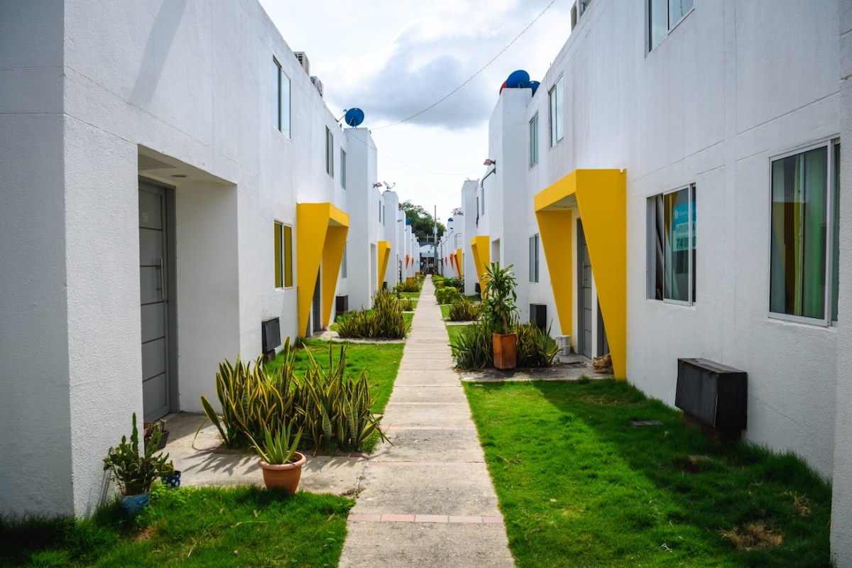 A wide pathway is bordered by white duplex houses featuring yellow accents. Lush greenery and potted plants are placed along the sides of the walkway, which leads through the neat garden area, inviting guests to explore the surrounding environment.