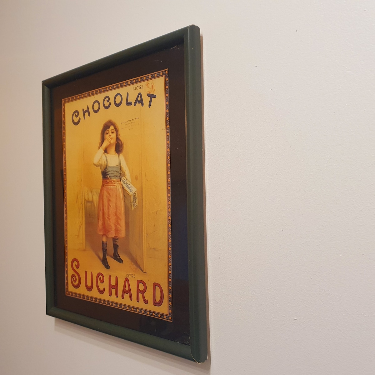 A vintage poster for Chocolat Suchard is displayed on a white wall. The artwork features a woman in period clothing, holding a chocolate bar. The border of the frame adds a touch of elegance, complementing the warm colors of the poster.