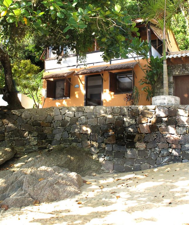 A cozy two-story house is nestled among lush greenery, with a warm orange exterior. The stone wall in the foreground leads to a sandy area, suggesting proximity to the beach. Large windows invite natural light into the living space, offering a view of the surrounding landscape.