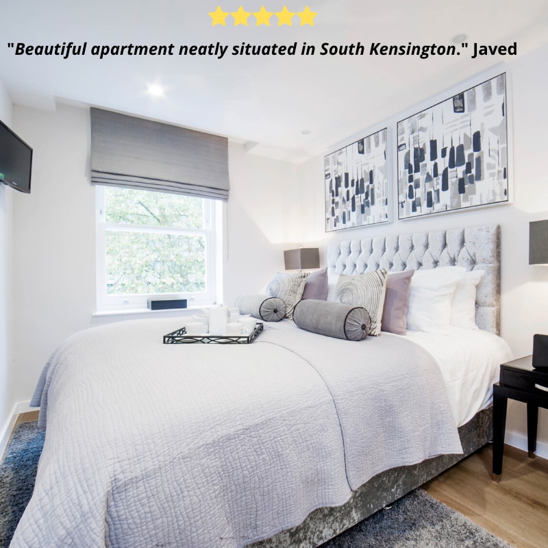 A well-appointed bedroom is featured with a spacious king size bed, adorned with soft grey bedding and cushions. Two pieces of abstract artwork hang above the headboard. A window provides natural light, partially covered by a light grey shade, enhancing the room’s inviting ambiance.