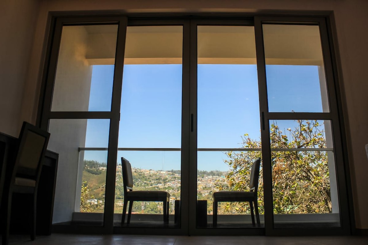 Two chairs are positioned thoughtfully on a balcony, facing an expansive view of the landscape beyond. Large glass doors frame the scene, allowing natural light to fill the space while creating a seamless connection between indoor and outdoor areas.