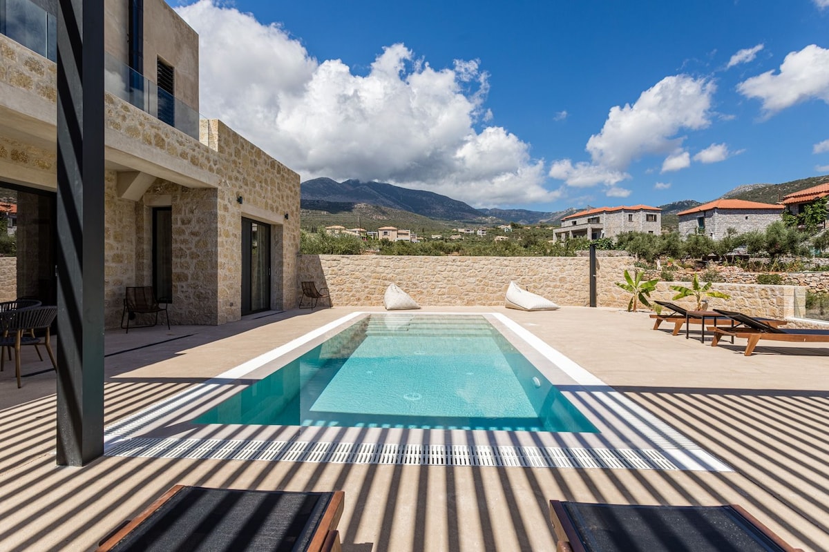 A tranquil private pool is surrounded by a spacious terrace, accented by sun loungers and natural stone walls. Lush greenery is visible in the background, with mountains and a partly cloudy sky creating a serene atmosphere. The sleek design of the villa adds to the inviting space.
