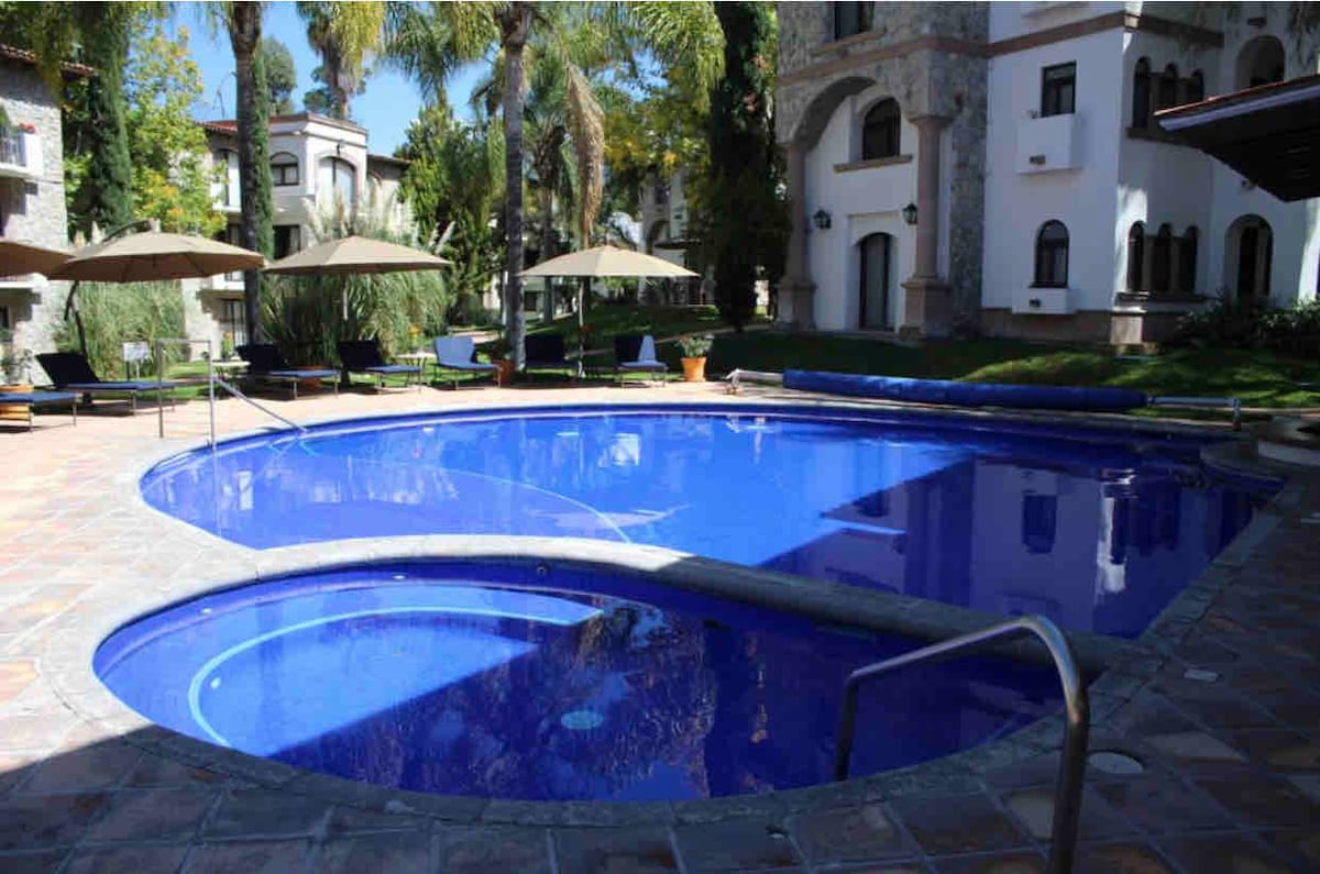 A spacious outdoor pool is framed by lounge chairs and shaded umbrellas. Lush greenery and palm trees surround the pool area, providing a serene environment. The pool features a unique shape with steps leading into the inviting blue water, creating a relaxing atmosphere.
