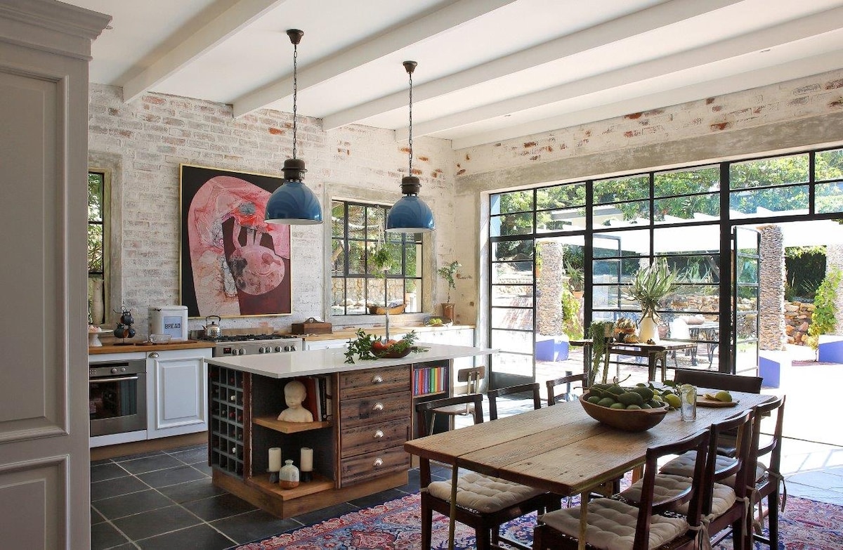 An open-plan kitchen and dining area is showcased with large glass doors, allowing ample natural light. Exposed brick walls add character. A central island with seating is surrounded by a wooden dining table and an eclectic selection of chairs. Decorative art and vibrant greenery are visible.