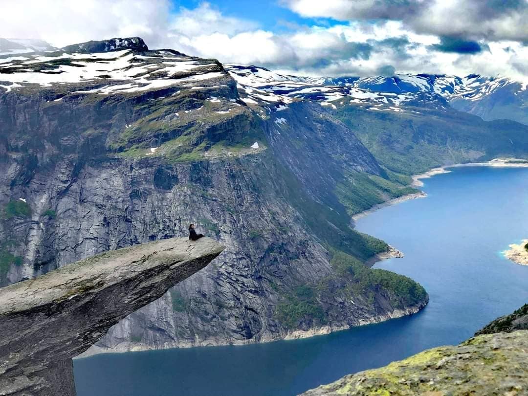 A stunning landscape shows a person sitting on a prominent rock formation at the edge of a cliff. Below, a fjord winds through lush green mountains, adorned with snow-capped peaks under a cloudy sky, creating a dramatic view of nature's beauty.