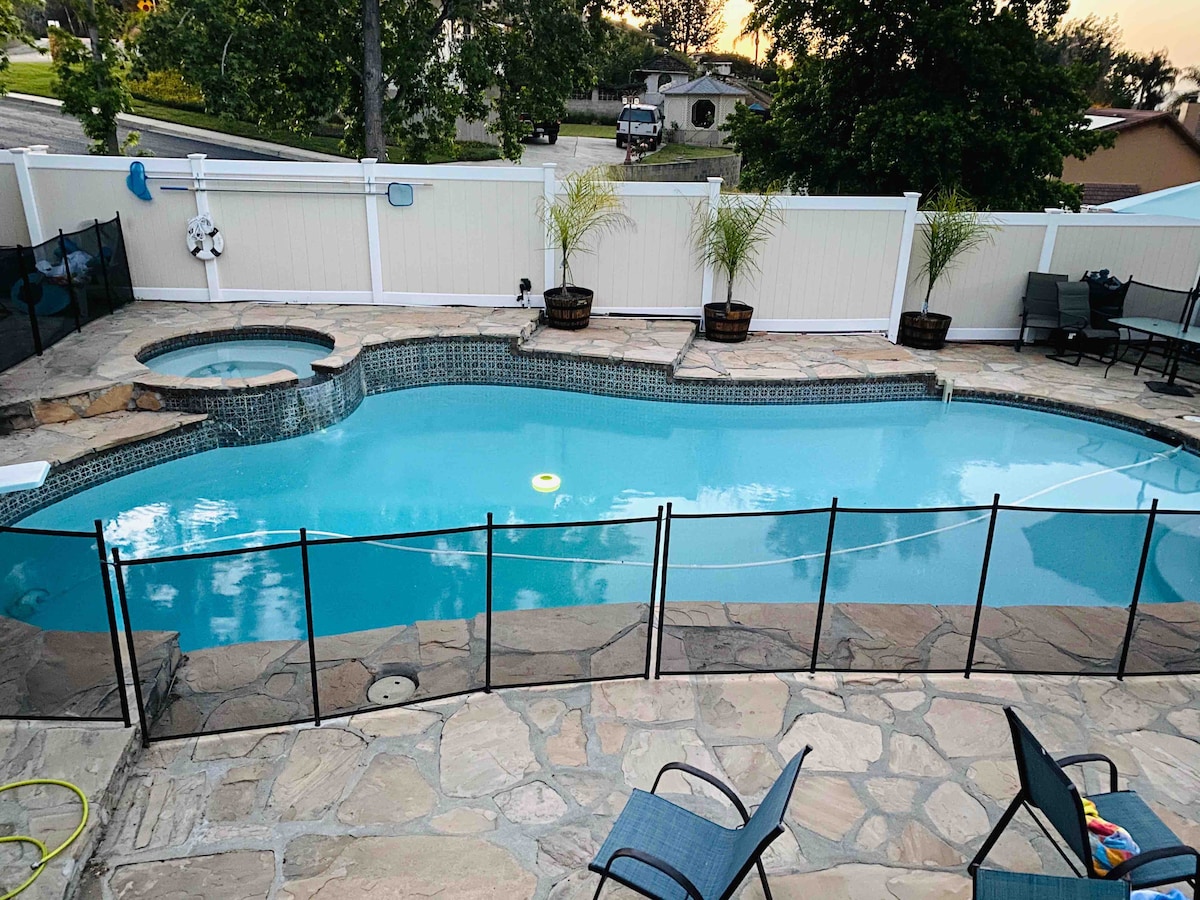 A serene saltwater pool is featured, surrounded by a stone patio and secured with a safety fence. A spa is integrated at one end of the pool, with two blue lounge chairs positioned nearby. Lush greenery is visible in planters along the patio.