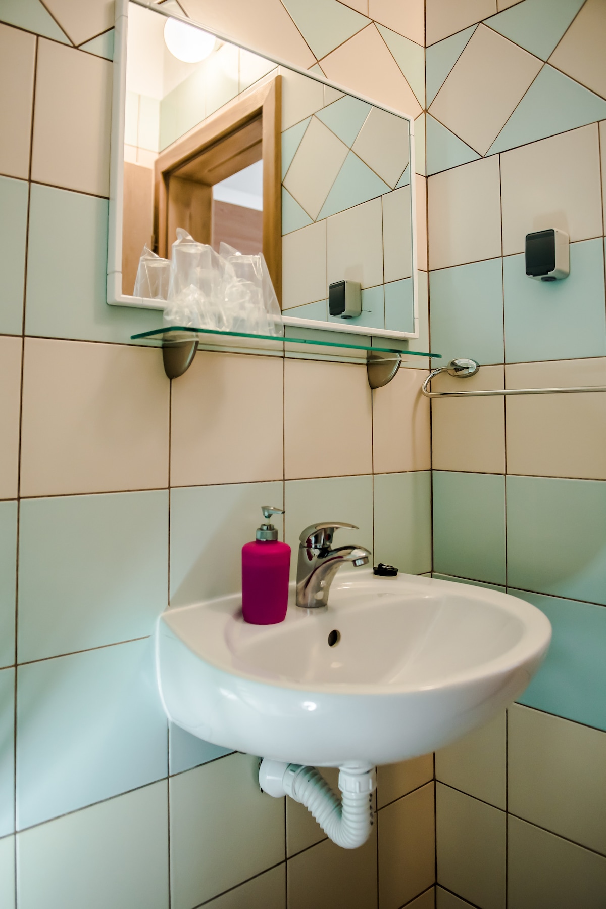 A compact bathroom is fitted with a wall-mounted sink and a mirror above it. The walls are adorned with geometric tiles in soft pastel colors. A shelf is mounted above the sink, displaying organized toiletries, while a liquid soap dispenser is positioned beside the sink.