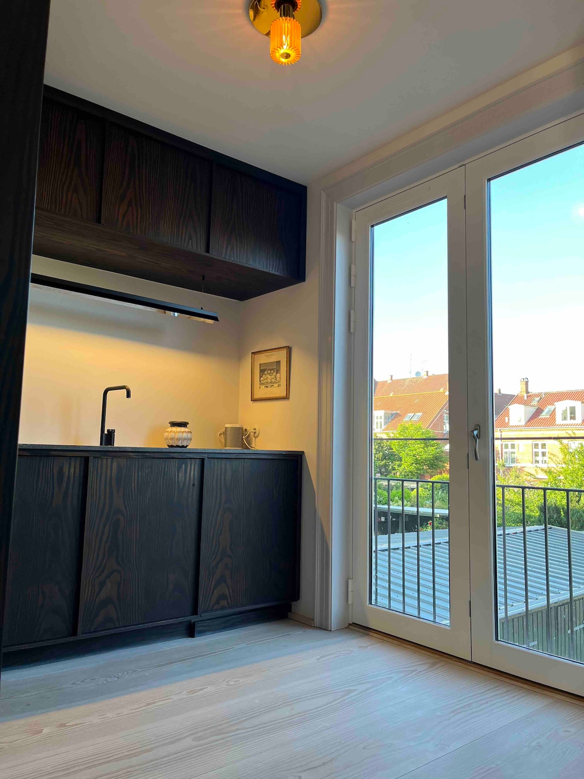 A compact kitchen space features dark cabinetry and a minimalist design. A modern faucet is visible alongside a jar and cup on the countertop. Large glass doors open to a balcony, allowing natural light to illuminate the area and offering views of the surrounding neighborhood.