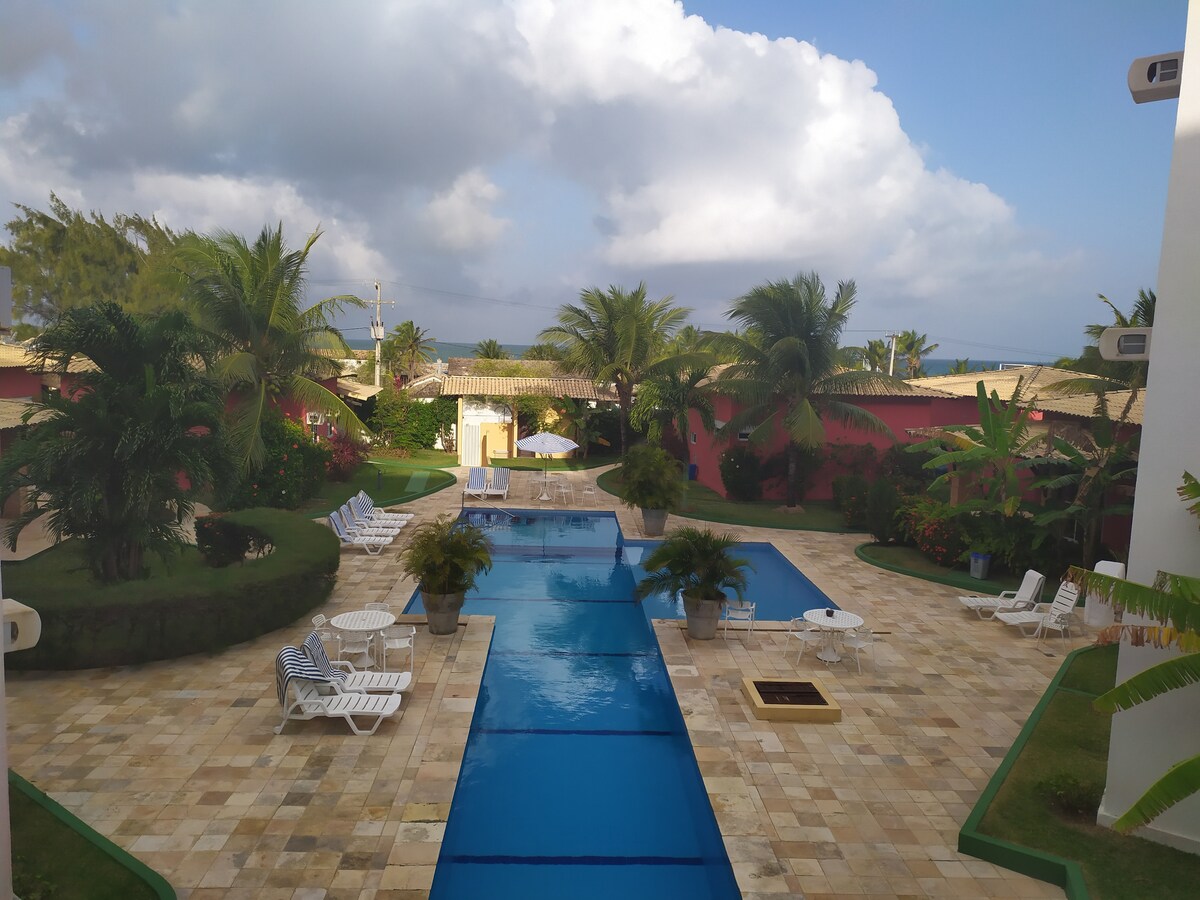 The outdoor area features a long, rectangular swimming pool surrounded by lush greenery and palm trees. Light-colored lounge chairs are arranged along the poolside, while pathways of natural stone create a welcoming atmosphere. A clear sky is visible above with scattered clouds.