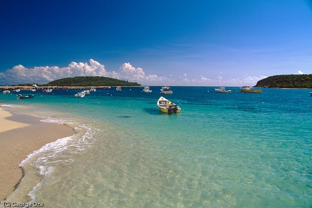 Calm turquoise waters gently lap against a sandy shoreline. Small boats are anchored nearby, while distant hills provide a natural backdrop under a bright blue sky dotted with soft white clouds.
