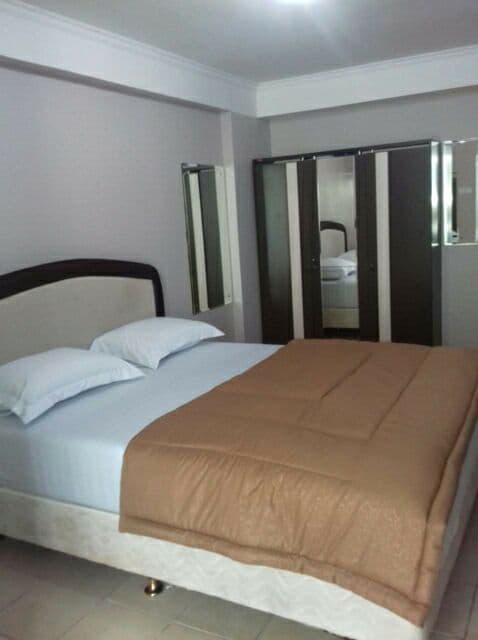 A furnished bedroom is presented, featuring a spacious bed with a light fabric headboard and a neatly arranged brown blanket. Mirrored wardrobes are positioned against one wall, reflecting the room's soft, neutral tones.