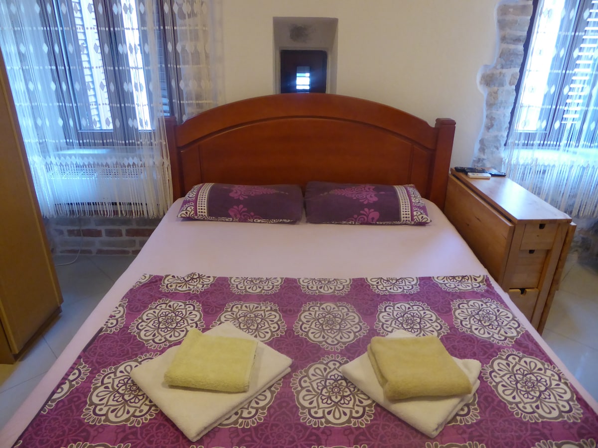 A comfortable sleeping area features a spacious bed with a purple patterned blanket. Two neatly arranged towels are placed on the bed, alongside a wooden nightstand. Light filters through the nearby windows, adorned with sheer curtains, creating a bright and airy ambiance.