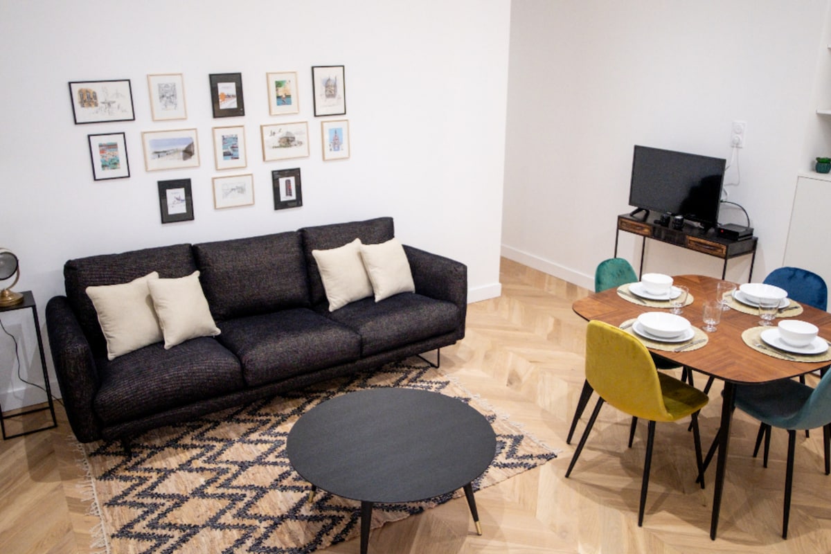 A spacious living area features a dark upholstered sofa adorned with neutral cushions. A round black coffee table sits atop a patterned rug, while a wooden dining table is set for four with colorful chairs. Framed art pieces embellish the white walls.