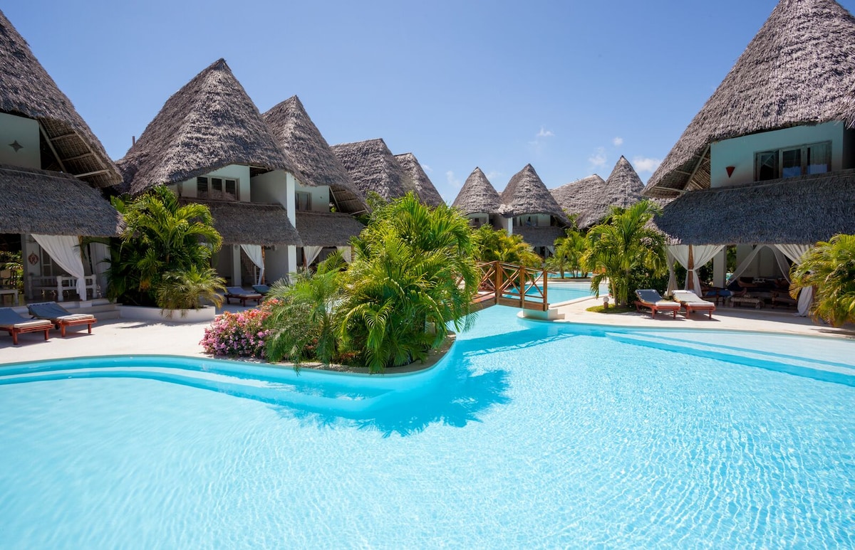 The image displays a clear blue swimming pool surrounded by lush tropical greenery and thatched-roof villas. Comfortable lounge chairs are positioned nearby, inviting relaxation. The bright sky and sunlight reflect on the water's surface, highlighting the serene atmosphere of the villa complex.