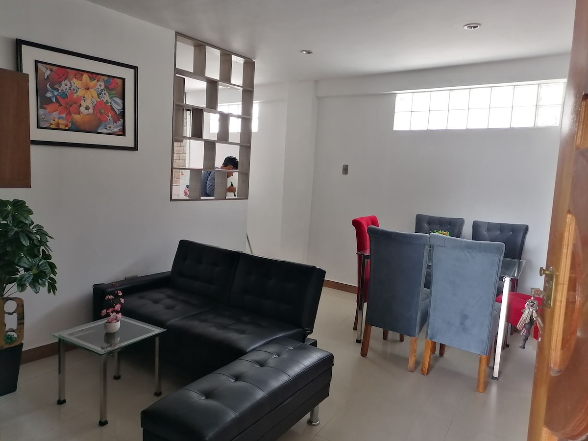 A modern living area features a black sectional sofa and a glass-top coffee table. Bright natural light enters through a window, illuminating a dining table with grey and red chairs. A decorative plant adds a touch of greenery to the space.
