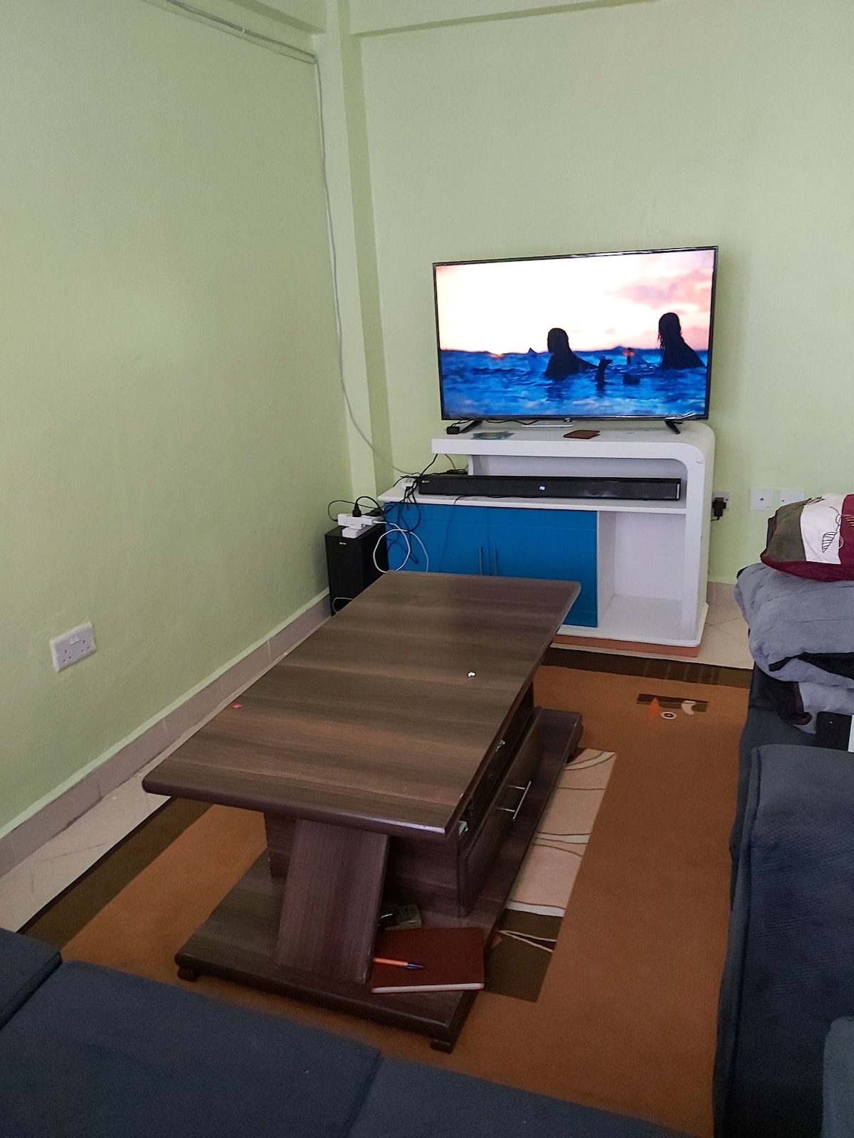 A compact living space is displayed with a small, dark wooden coffee table at the center. A television mounted on the wall presents a scenic image. A blue TV stand is positioned below, and a gray couch is visible in the background, complemented by an assortment of miscellaneous items.