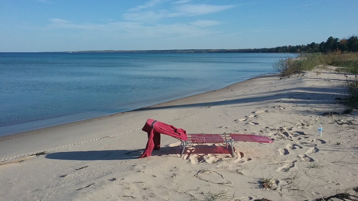 A sandy beach stretches along the calm water of Lake Huron, with a red lounge chair positioned on the shore. Gentle ripples create a serene surface, while sparse vegetation lines the beach, indicating a tranquil setting for relaxation.