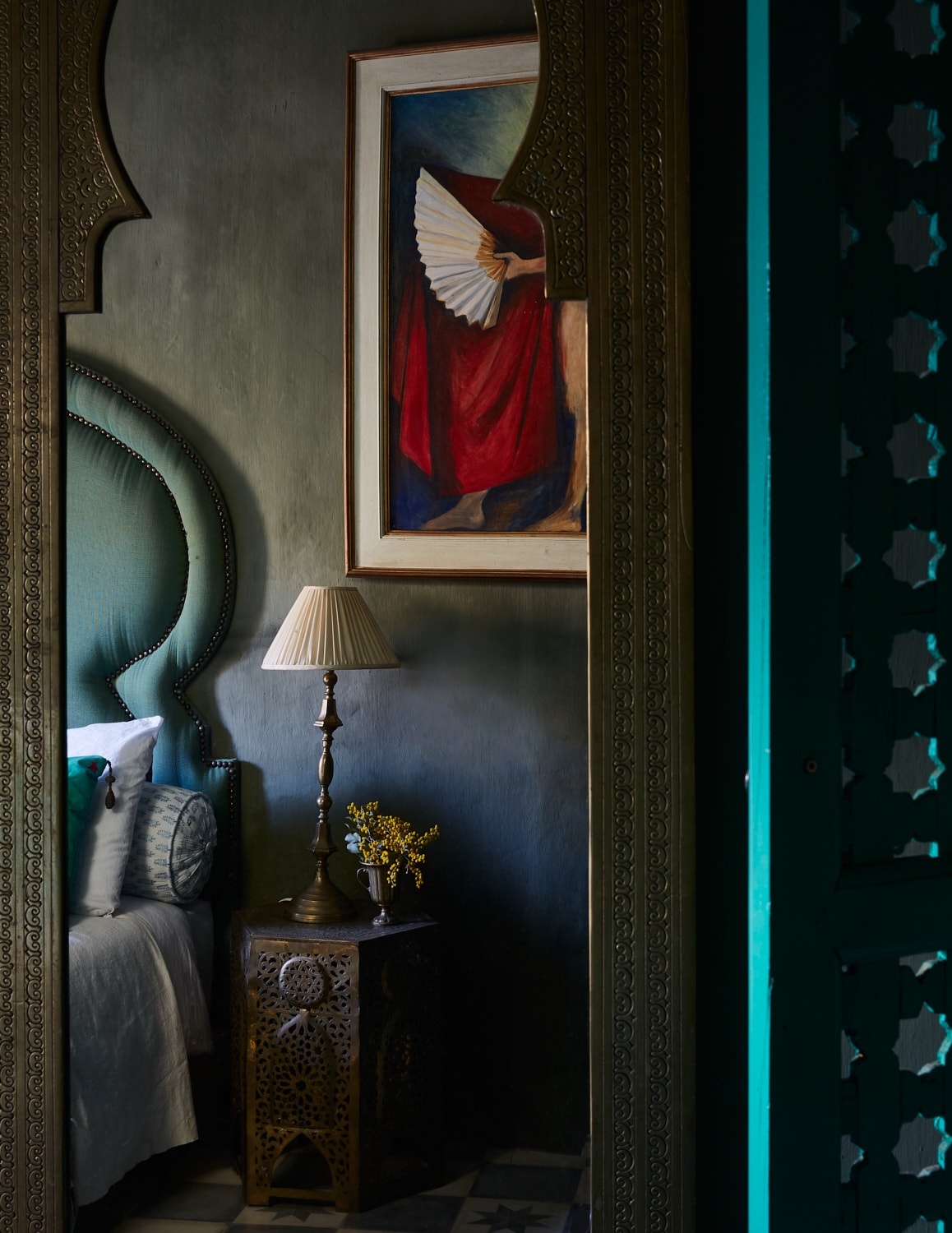 A softly lit bedroom is framed by a decorative mirror, highlighting a tall headboard and a bedside table with a lamp. A colorful painting adorns the wall, while a small vase of flowers adds a touch of freshness. Intricate patterns are visible on the nearby door.
