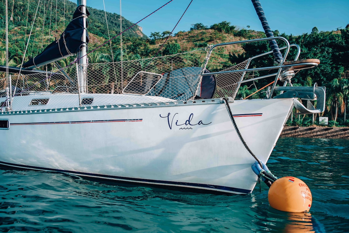 The bow of the sailboat 'Vida' is captured, displaying its sleek hull and the name elegantly painted. It is anchored with a buoy nearby, surrounded by calm waters and lush greenery from the shoreline in the background.