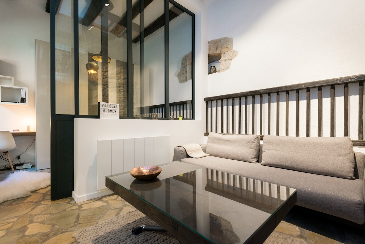 A modern living area features a comfortable gray sofa positioned near a sleek glass coffee table. The space is illuminated by warm lighting, with a unique decorative bowl resting on the table. The walls showcase a blend of textures, enhancing the room's character.