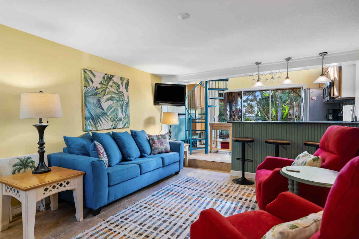 A living area with a blue sofa and two red armchairs is complemented by a decorative area rug. A tropical-themed wall art piece adds a splash of color. A staircase leads to the upper level, with a dining area visible in the background.