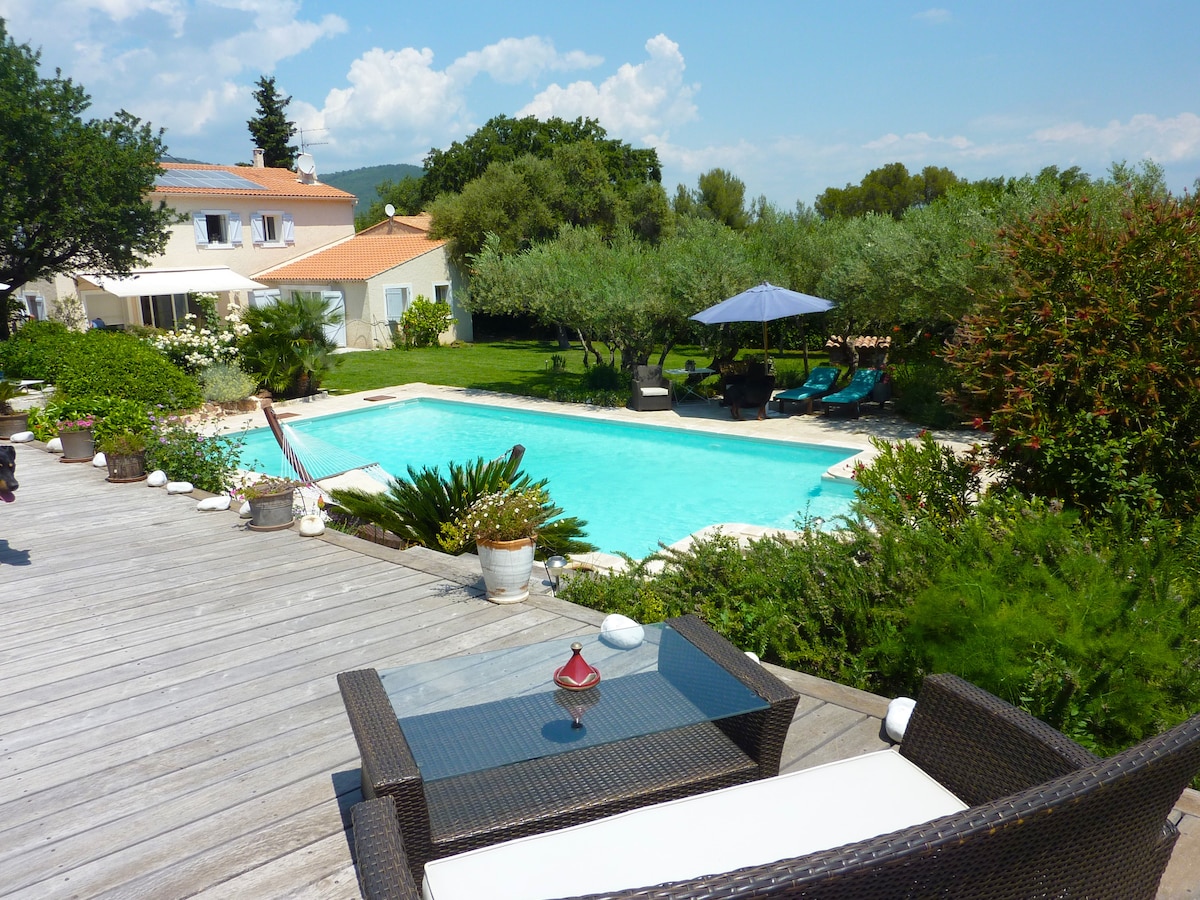 A view of the shared pool surrounded by greenery and sun loungers. The pool's turquoise water reflects the blue sky. A wooden terrace features a glass-top table with seating, positioned among potted plants, providing a relaxing outdoor space.