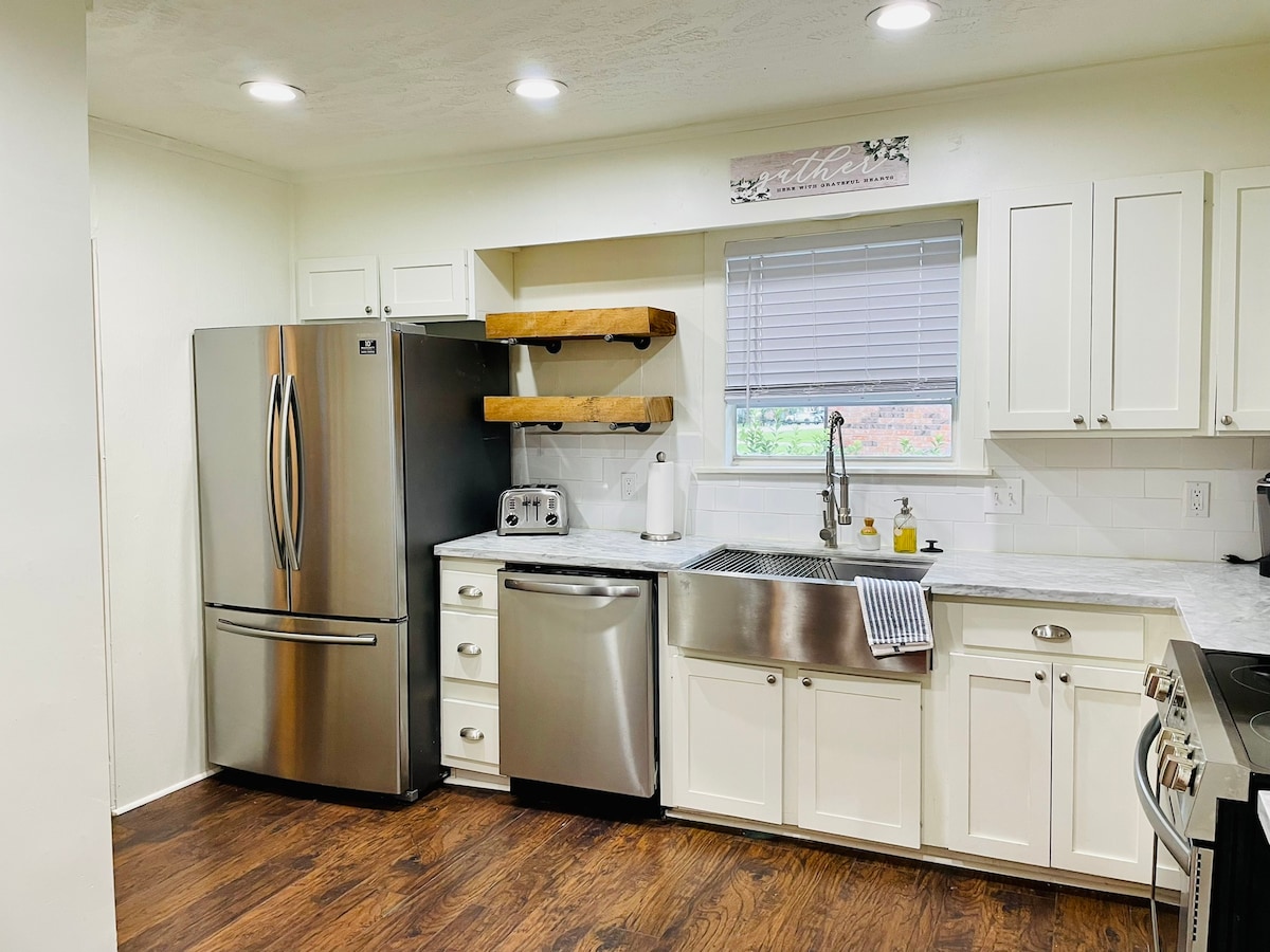 The kitchen features stainless steel appliances, including a refrigerator and dishwasher. Open shelving displays kitchen essentials, while a stainless steel sink is set against a window that allows natural light. White cabinetry and warm wood flooring complete the functional space.