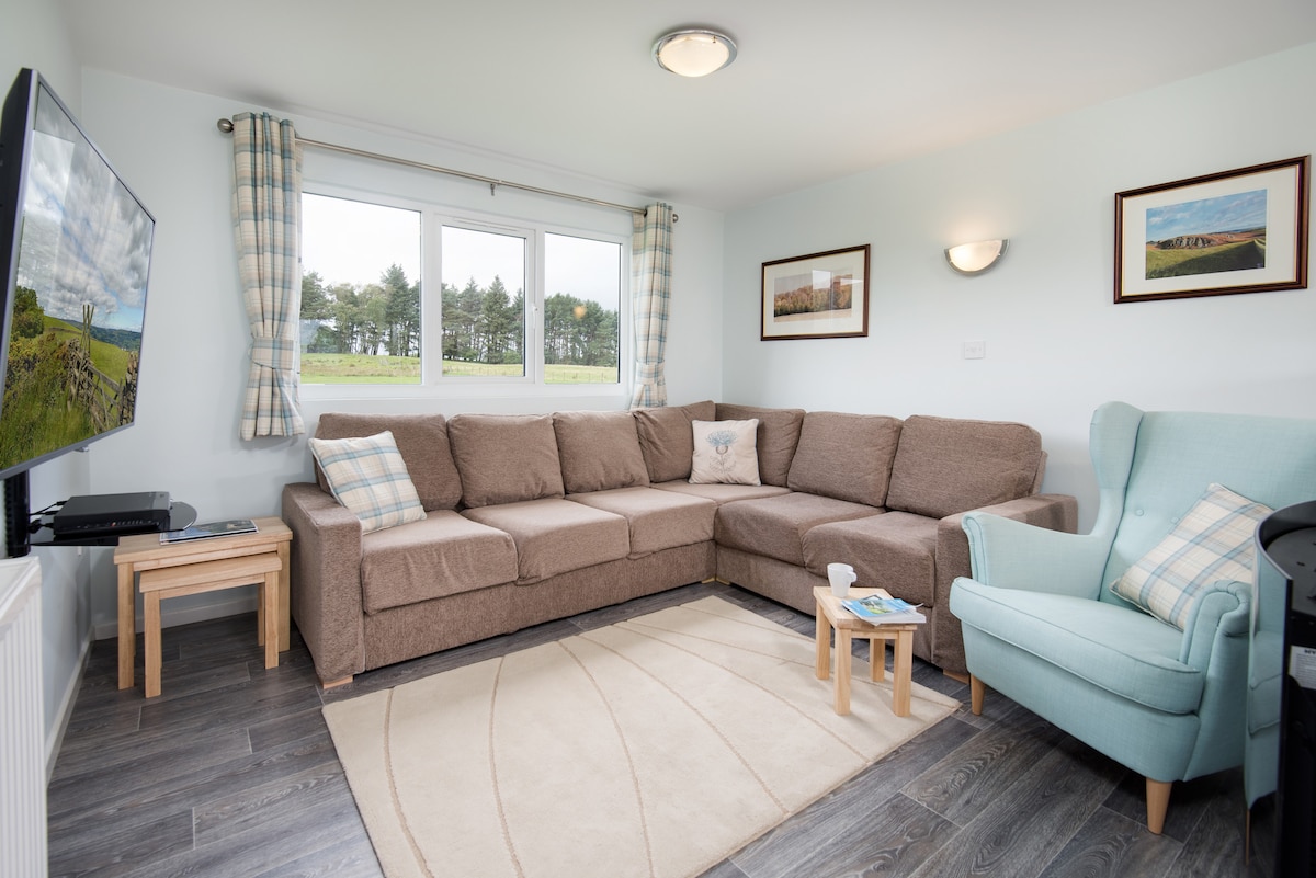 A spacious lounge area features a large, U-shaped sofa in a neutral tone, accompanied by a light blue armchair. A small wooden table sits in front of the sofa. Large windows offer views of the surrounding countryside, while a wall-mounted flat-screen TV is positioned nearby.