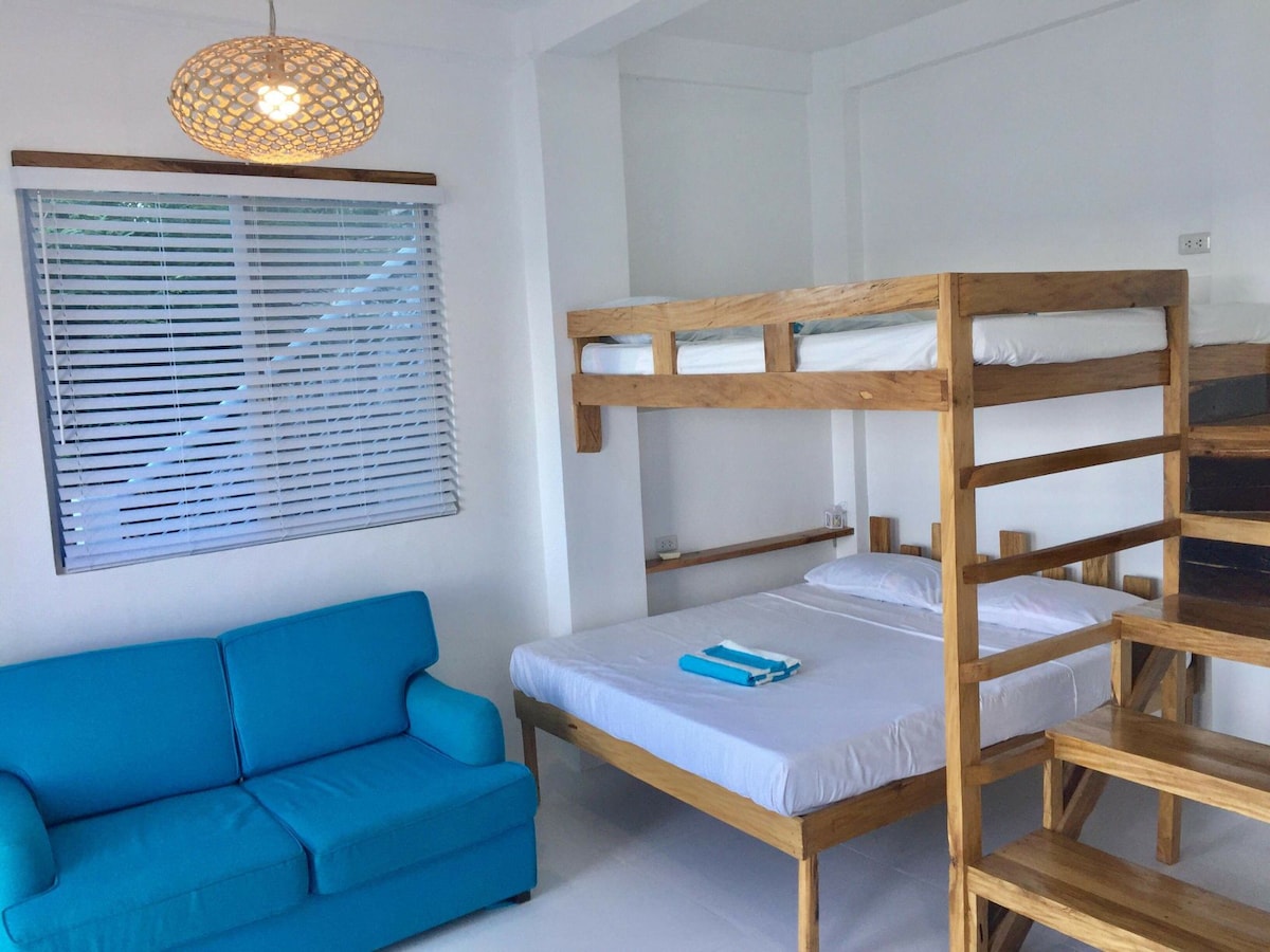 A bright and airy room features a double-decker wooden bed with two queen-size mattresses on the upper and lower levels. A comfortable blue sofa complements the space, which is enhanced by natural light filtered through the window with horizontal blinds.