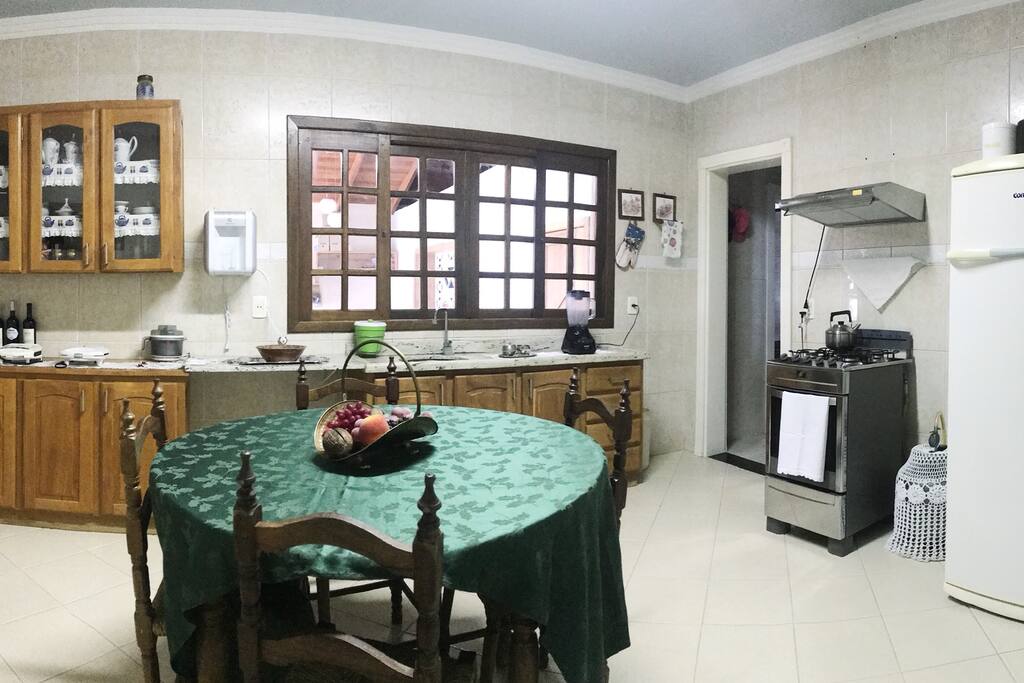 Cozinha grande com fogão elétrico e exaustor, geladeira, armários, pia, água filtrada, fogão de chapa à gás (semelhante ao fogão à lenha), mesa para 4-6 lugares.