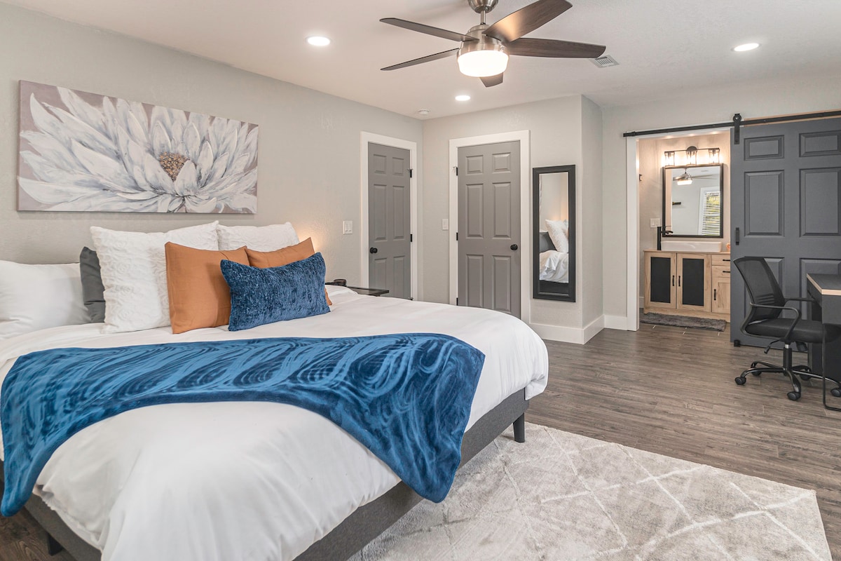 A spacious bedroom is highlighted by a large bed featuring white linens and decorative pillows in shades of blue and orange. A ceiling fan circulates air, while a work desk and chair are positioned nearby. The room is warmly illuminated and equipped with subtle decor.