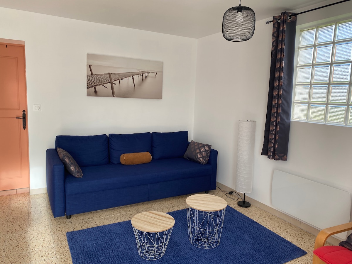 A living area features a blue sofa accompanied by a cushion and two small wooden tables. Natural light enters through a glass block window, enhancing the space. A floor lamp and a wall-mounted artwork of a pier complete the room's minimalist design.