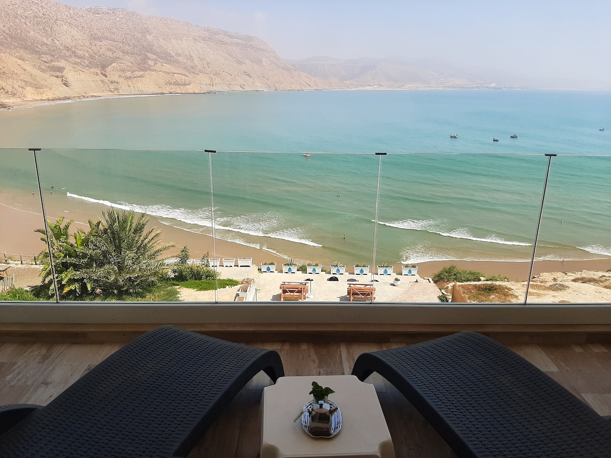 A serene view of the Atlantic Ocean is presented from lounge chairs, featuring a glass railing that provides an unobstructed perspective of the beach. The landscape includes golden sand, gentle waves, and a backdrop of mountains under a clear sky.