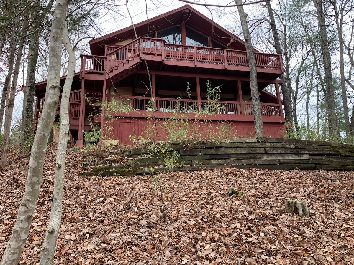 The exterior of a two-story red wooden house is framed by trees and natural foliage. Multiple decks are visible, showcasing railings and access points. The ground is covered in fallen leaves, creating a rustic setting amidst a peaceful wooded area.