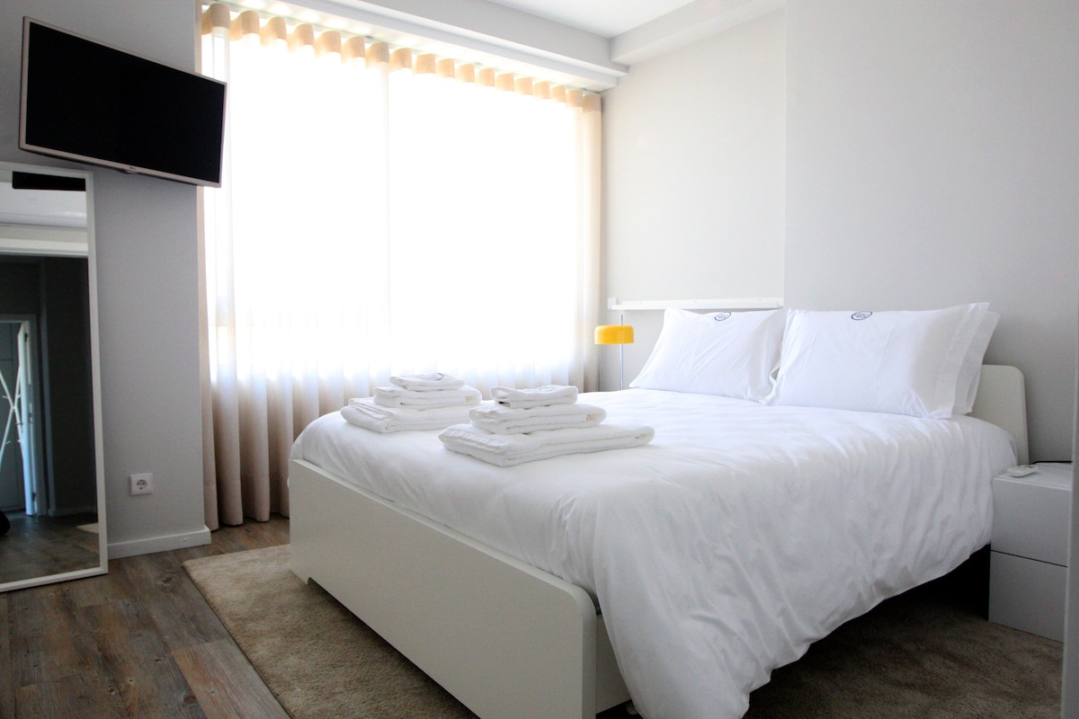 A bright sleeping area is featured, showcasing a spacious bed neatly made with white linens. Soft natural light enters through large windows adorned with sheer curtains. A flat-screen TV is mounted on the wall, and a full-length mirror stands nearby, reflecting the inviting surroundings.