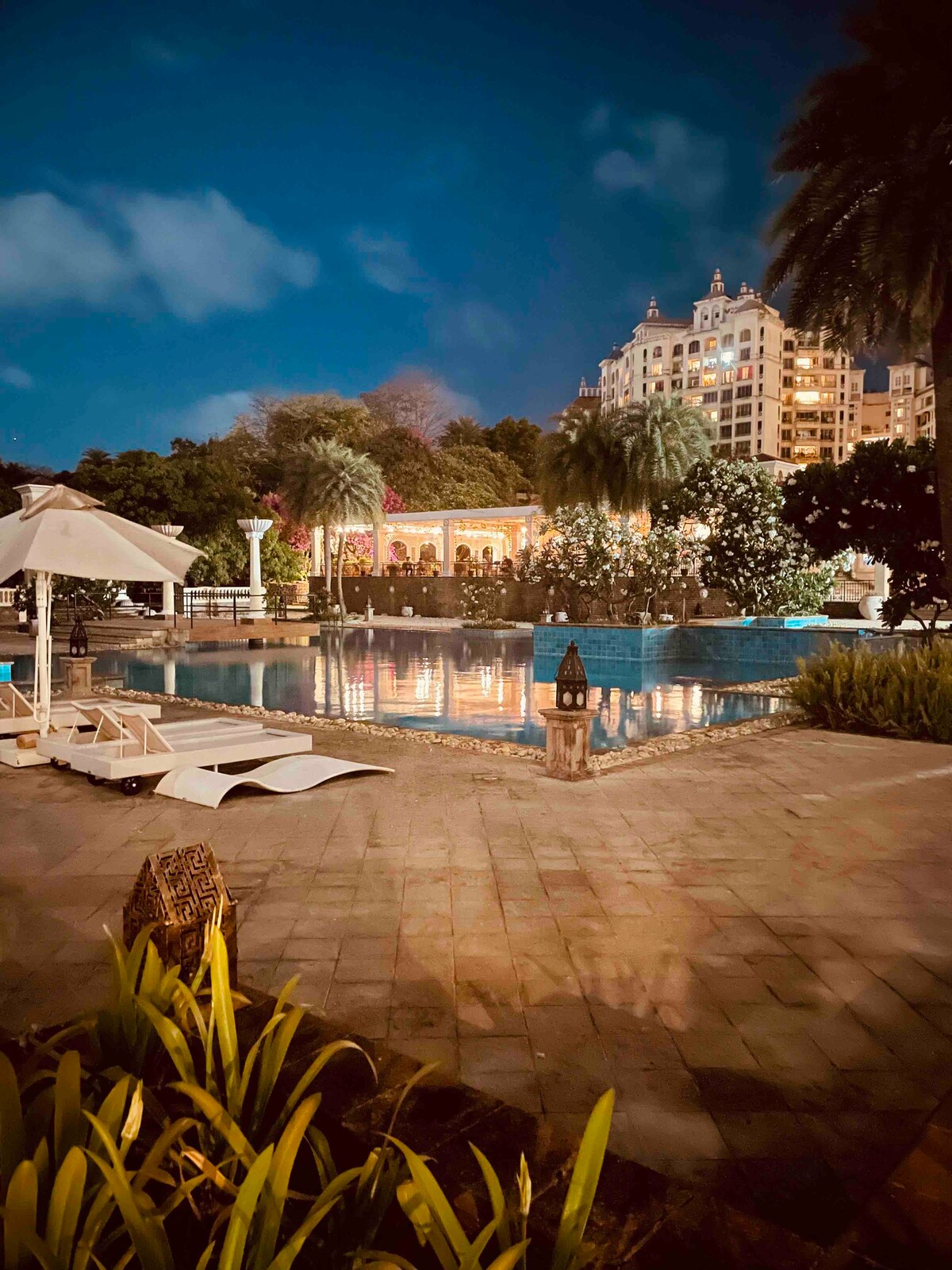 Evening lights illuminate a serene swimming pool surrounded by tropical foliage and elegant loungers. The well-maintained patio features ornate decor elements, while palm trees sway gently nearby. A luxurious building emerges in the background, enhancing the tranquil setting.