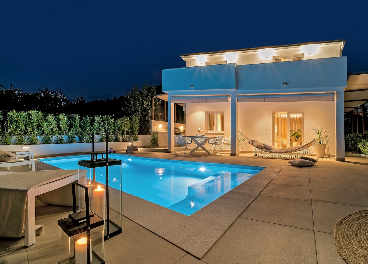 The image showcases a modern house illuminated by warm lights against a night sky. A tranquil swimming pool reflects the surroundings, while a hammock and seating area invite relaxation. Soft landscaping adds a touch of greenery, complementing the well-lit outdoor dining space.