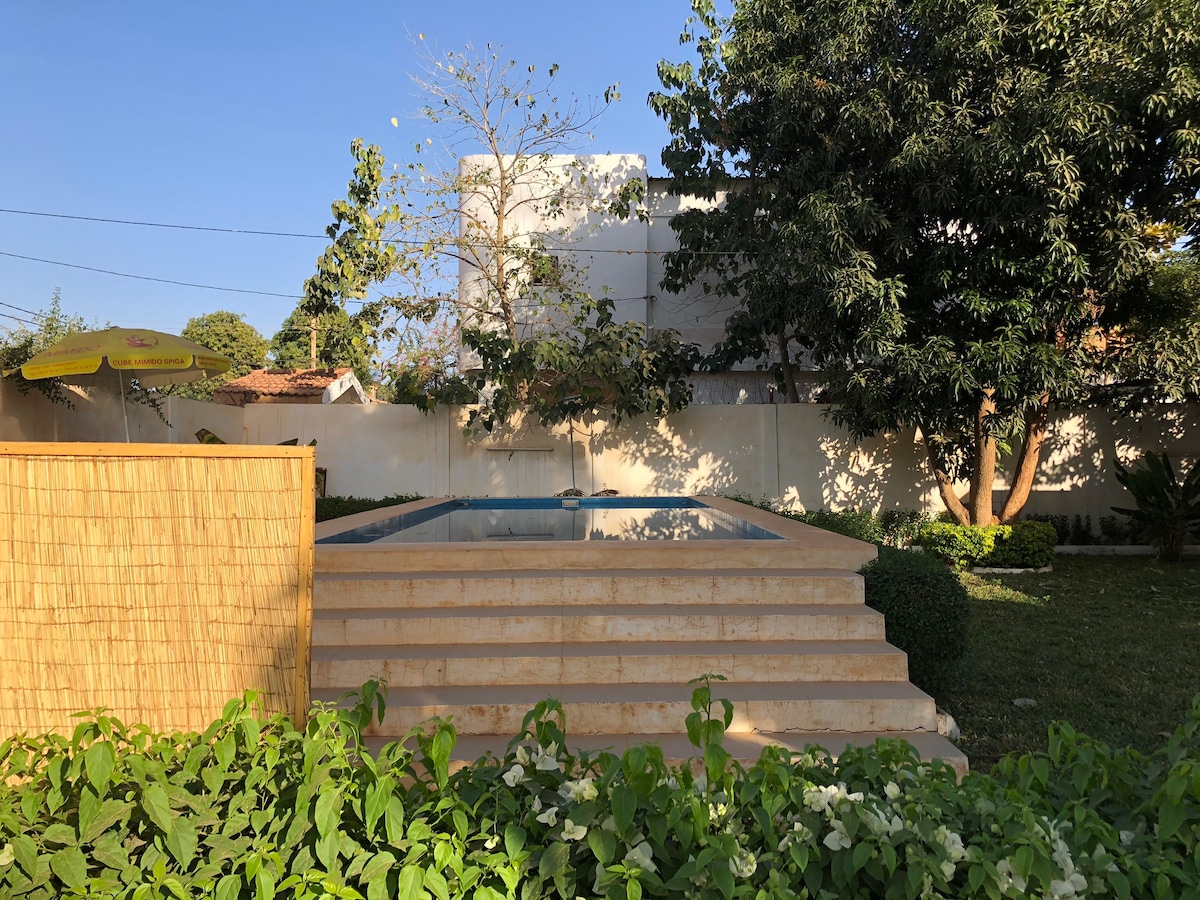A serene garden area is surrounded by lush greenery and features a swimming pool, elevated with tiered steps leading to its edge. Various plants and trees provide privacy, while a shaded area is visible nearby, offering a comfortable spot to relax.
