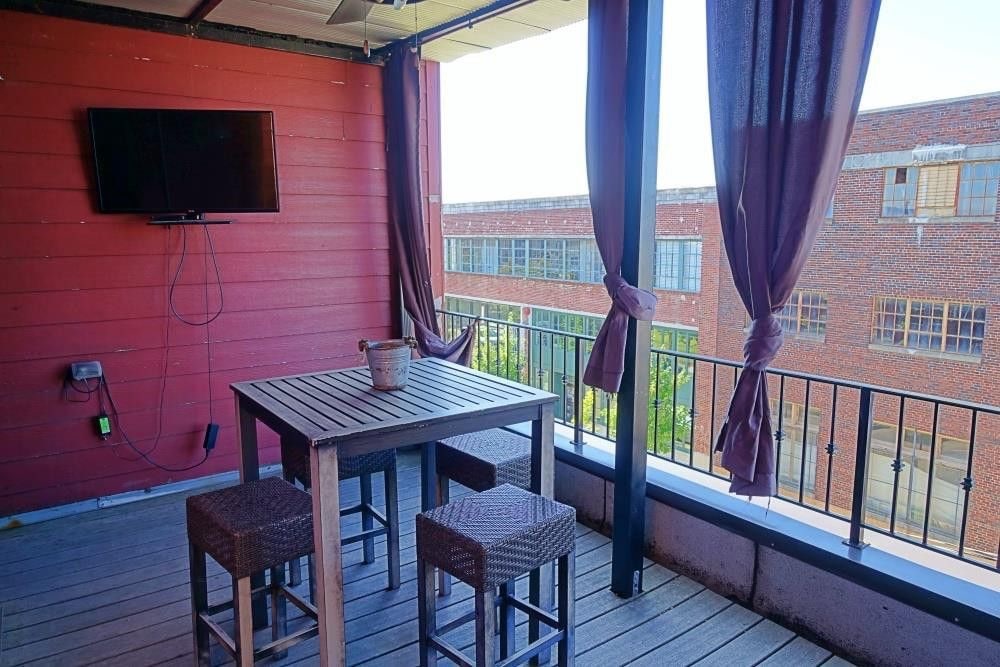 A private patio with a wooden table and four high stools is shown. Sheer drapes frame the space, providing some shade. A wall-mounted television is visible, enhancing the outdoor experience. The backdrop includes an industrial-style building with large windows.