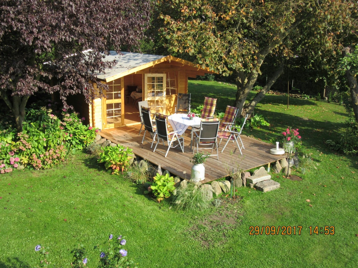 A wooden gazebo is set on a wooden deck surrounded by lush greenery. A table is arranged for outdoor dining, complemented by chairs with cushions. Flowering plants and tall trees provide a natural border, creating a serene outdoor space perfect for relaxation.