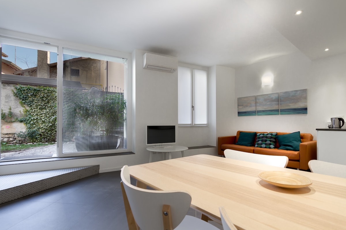 A light-filled living area features a sofa with colorful cushions and a flat-screen television on a modern stand. A dining table with white chairs is positioned nearby, and large windows provide views of an outdoor space with greenery and a patio.