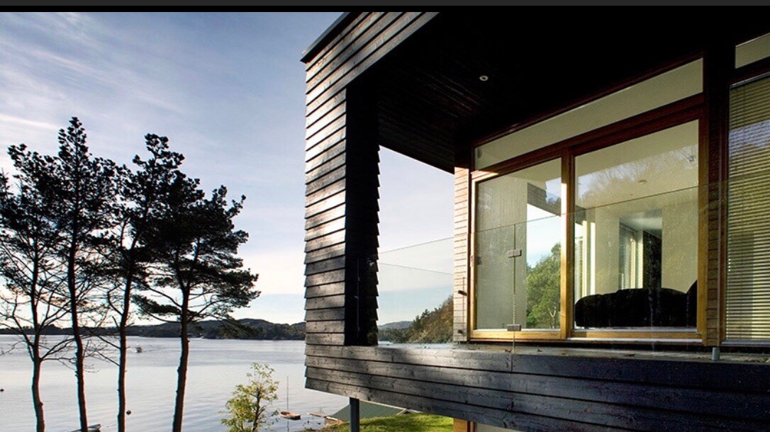 The image presents a modern villa with large glass windows overlooking a serene body of water. Features include a sleek wooden exterior and trees framing the view, with the reflection of the house visible on the water's surface.